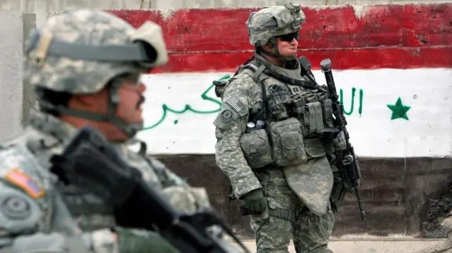 两名身著作战装备的美国士兵站在一块涂有伊拉克国旗颜色的路障旁，一名士兵在前景，另一名士兵手持步枪在背景。