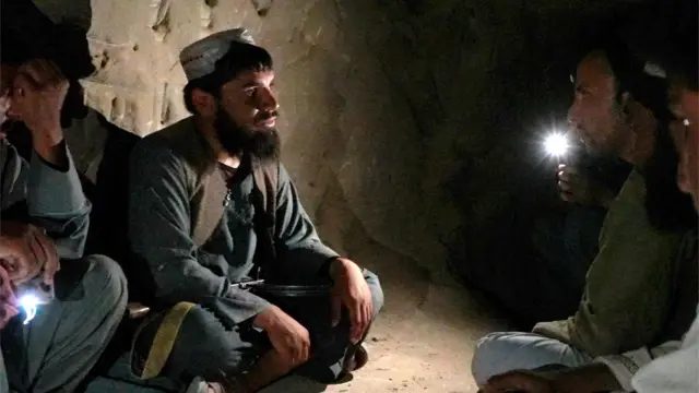 离婚、智能手机与管控——BBC观察塔利班如何治理阿富汗 6 一名留着胡须的男子身穿深色传统长衫和长裤,头戴无帽簷圆帽,盘腿坐在昏暗洞穴内的裸露地面上。他位于画面左侧,正看着坐在右侧的一名男子,后者手持手电筒。