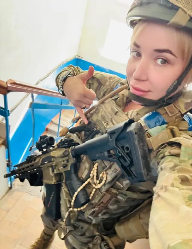 凯蒂·莱什卡谢利身穿作战服，胸前挂着狙击枪，站在一处室内的楼梯顶，右手在比手势