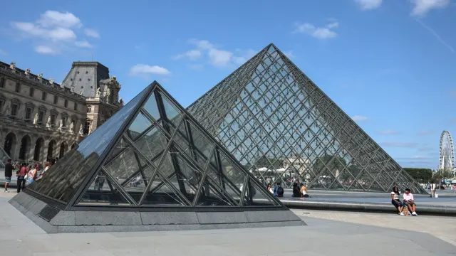 巴黎罗浮宫的两座现代玻璃金字塔，左侧则是一座较古老的建筑。天空湛蓝，金字塔前坐着几个人。远处右侧可以看到一座摩天轮。 