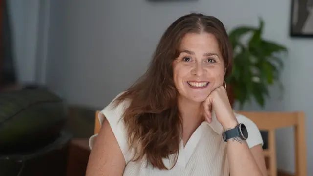 Jessica Hill 是一名30多岁女性，留着长长的红棕色头发。她手托下巴，对着镜头微笑。她穿着白色短袖有领衬衫，手腕上戴着 Garmin 智慧手表。她坐在木椅上，背景可见一株大型室内植物，但呈失焦状态。