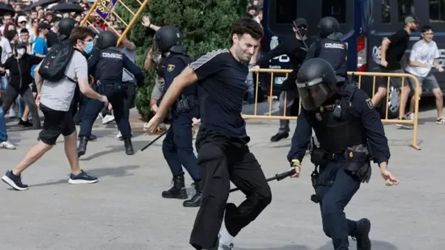 一名防暴警察似乎正用警棍抵着一名抗议者的腿后侧。背景中可以看到成群的抗议者，以及几名身穿制服、头戴头盔的防暴警察。远处，有人正在抬起一段黄色围篱。