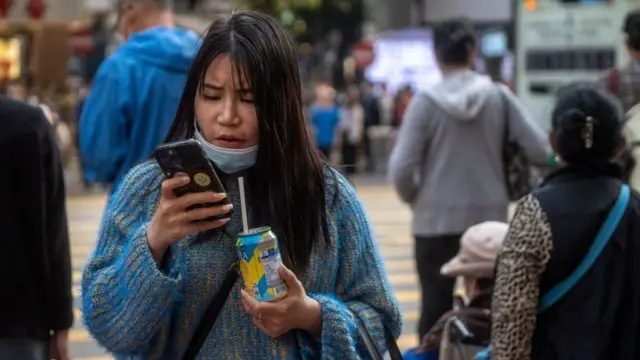 一名女子在街上看着手机，拿着一罐饮料，她的口罩拉至下巴位置。