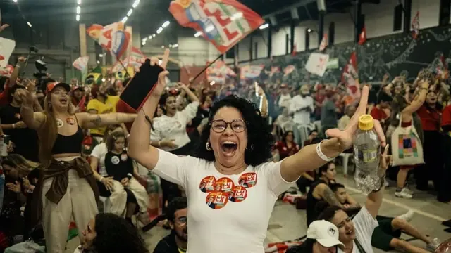 巴西圣保罗，在选后集会上，卢拉的一位支持者在选举结果公布时做出反应，背景中各党派的旗帜迎风飘扬。她穿着一件贴满卢拉头像贴纸的白色上衣。（25/10/2026）
