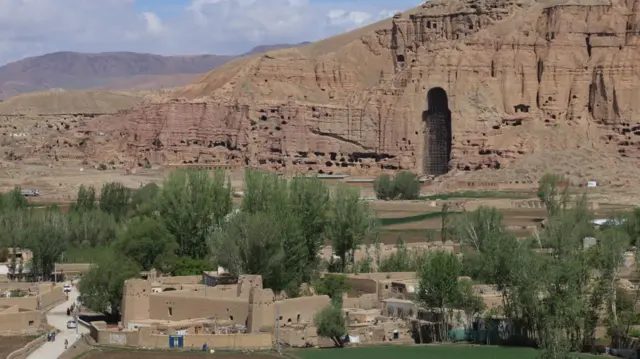 2024年拍摄于阿富汗巴米扬山坡上的巨大洞穴，该处曾矗立一座巨型佛像。洞穴位于裸露、泛红色岩石构成的悬崖之中，前景可见树木和低层建筑。