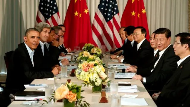 习近平（右二）与奥巴马（左一）在加州兰乔米拉市安纳伯格庄园连同两国官员举行会晤，长桌上摆满玫瑰花盆与水杯，后方有两国翻译和中美两国国旗（8/6/2013）