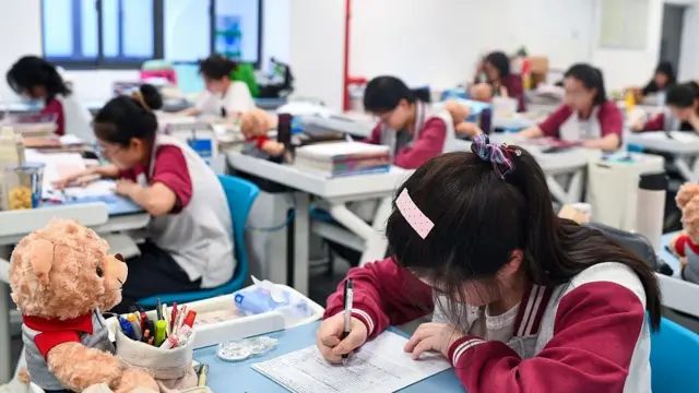课室内，一名青少年坐在书桌前低头看着桌上的纸张，并用黑色笔在纸上书写。书桌上放有文具和一个玩具熊。背景可见多名青少年分散坐在各自的书桌前，身处同一课室环境。
