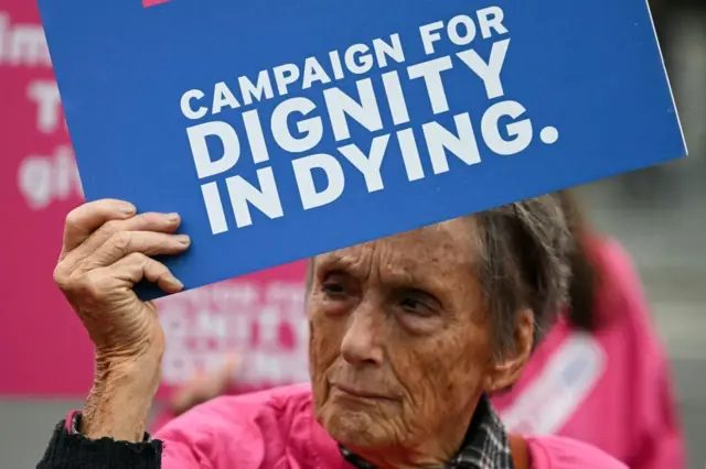 “尊严死亡”（Dignity in Dying）组织的一名倡议人士在英国国会所在地西敏宫外举行的示威活动中手持标语牌（16/10/2024）