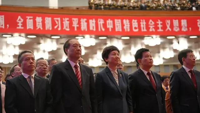 着正装的一排人站在人民大会堂里，背景挂着横幅“全面贯彻习近平新时代中国特色社会主义思想”