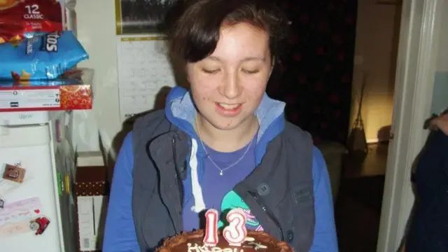 加比低头看着一个巧克力生日蛋糕，上面写着数字13。她穿着一件紫色上衣和一件海军蓝背心。