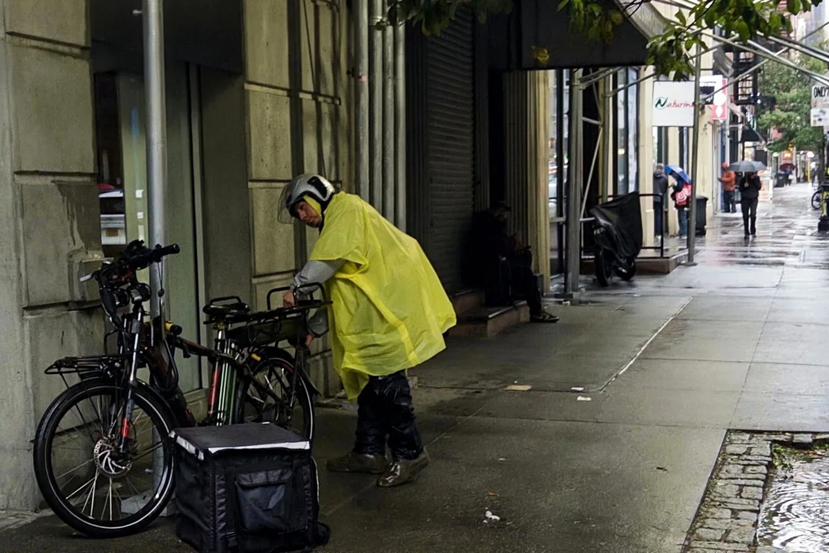 雨天的人行道上，一名穿黄色雨衣、戴头盔的外卖员站在电动自行车旁，地上放着外卖保温包