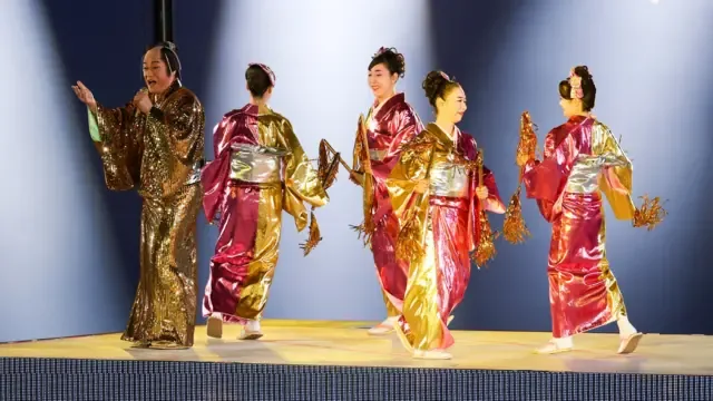 2026年10月4日，名古屋瑞穗公园陆上竞技场，身穿金色和服的日本演员兼歌手松平健（左）于爱知—名古屋亚运会闭幕式上演唱，身旁有四名身穿闪亮粉金色和服的女子伴舞