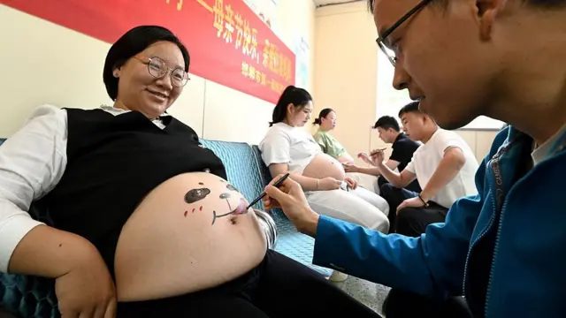 一名孕妇坐着露出腹部，一名男子正在她的肚皮上画图，背景亦有其他孕妇和陪同者。