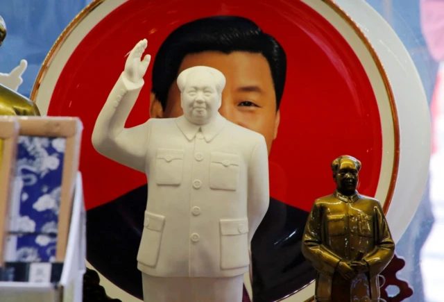 两个毛泽东雕像摆放在一个装饰盘前，盘子上画着微笑的习近平，其中较大的雕像遮住了习近平的大部分脸。