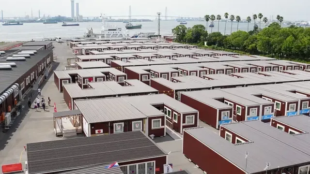 2026年9月18日，从高空俯瞰，可见位于日本名古屋海滨的一大片住宿设施区。整个园区由多排棕色货柜式组合房组成，配有灰色屋顶，整齐地以网格状排列。各栋建筑之间设有笔直的人行通道和道路，少数行人正在其间步行。
