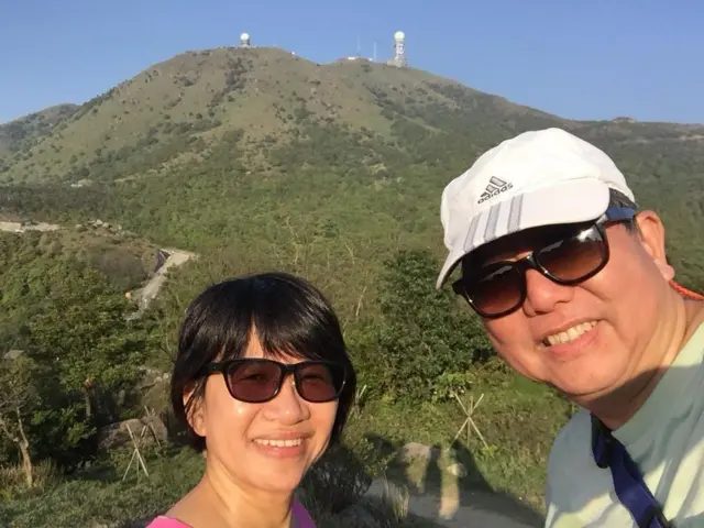 邓燕娥和李卓人在山上戴着墨镜拍照，李卓人戴着一顶白色帽。