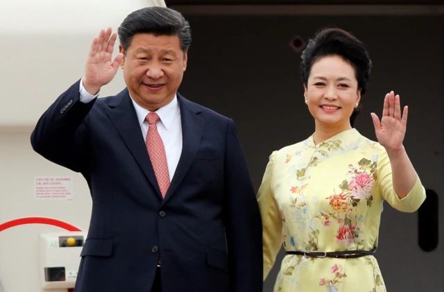 彭丽媛身穿碎花连身裙，与身穿西装打着红色领带的丈夫习近平在敞开的飞机舱门前挥手致意。 