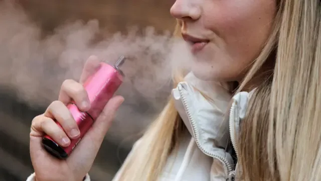 一名金发女子手持一支红色电子烟，吐出蒸气。
