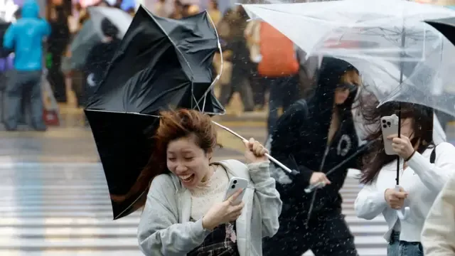 日本东京新宿街头，几位年轻女士在台风杜鹃的强风中举伞前行，雨伞被吹翻，略显狼狈（21/9/2026）