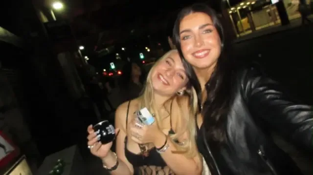 昏暗的酒吧里，两个女孩对着镜头微笑。左边的女孩是金发，穿着吊带背心，手里拿着一杯酒。她靠近左边拿着相机的朋友，两人一起自拍。右边的女孩留着棕色长发，穿着黑色皮夹克。