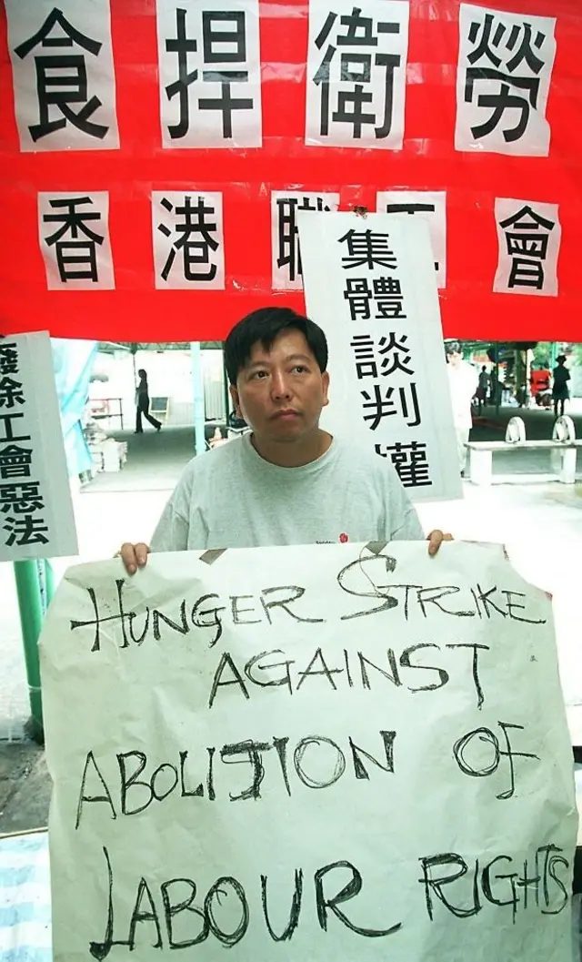 李卓人手持“HUNGER STRIKE AGAINST ABOLITION OF LABOUR RIGHTS”（
绝食抗议废除劳工权益）的横幅。