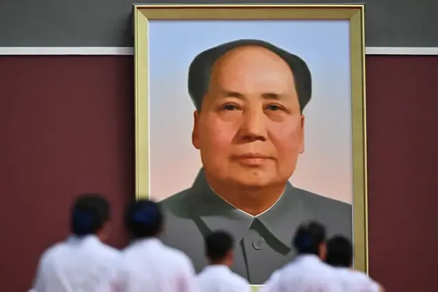 一幅大型毛泽东肖像悬挂在红色墙壁上，照片从下方拍摄，前景有数名身穿浅色衣服的人站立，面向肖像。