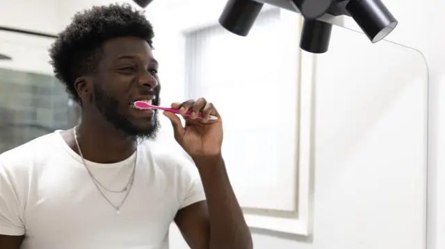 一个男人一边刷牙一边对着镜子。他穿着白色T卹，戴着银色项鍊。他的牙刷是粉红色的，他身后的浴室是白色的，淋浴间铺着灰色磁砖。