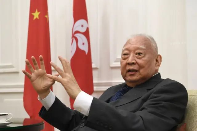 董建华坐在沙发上，背景是中国国旗和香港区旗