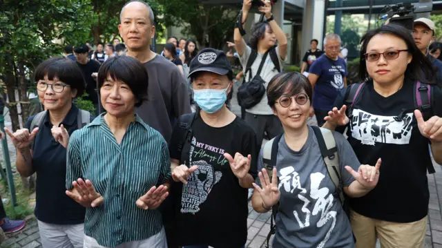 香港西九龙法院大楼外，邓燕娥（左二）、刘家仪（右二）、邹幸彤母亲刘华珍（中）、前社民连义工李盈姿（左一）、前支联会常委关振邦（后）与前泛民主派区议员陈剑琴（右一）举起“六四”手势合照（11/9/2026）
