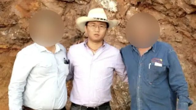 一名男子戴着白色牛仔帽，身穿浅紫色衬衫并将衣襬束进裤内。他戴着眼镜和项鍊，正望向镜头。他左侧及右侧各有一名男子，这两人的面容均经模糊处理，以隐藏其身份。